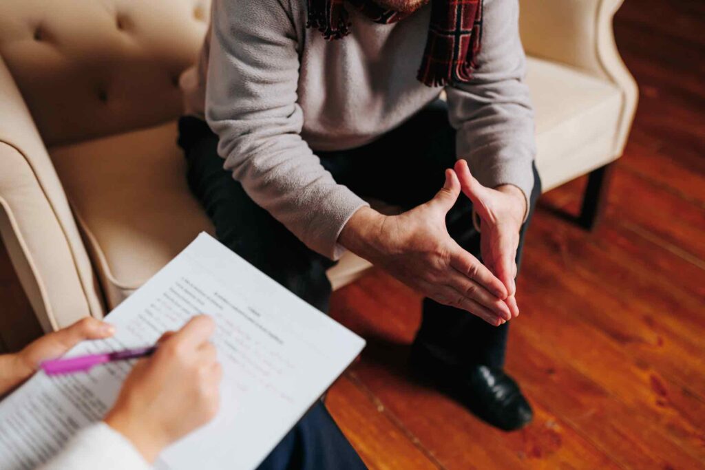 A counselor and client engage in a serious discussion. The client gestures expressively while seated, and the counselor takes notes with a pen on a clipboard.